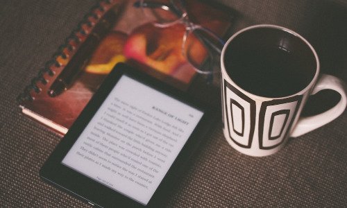 e-reader, coffee cup, glasses on top of notepad.
