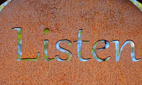 A rust colored sign with the word Listen carved in the center.