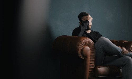 White man with brown hair and beard, sitting on a brown couch with his hand over his face in a distressed manner.