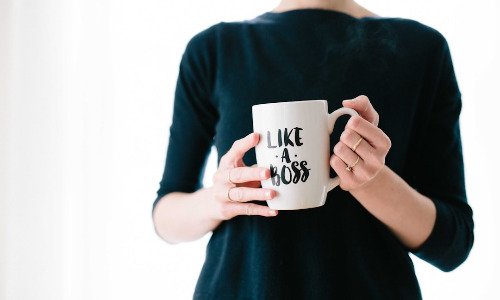 Hands holding a white mug with the words Like a Boss written in black cursive on it. The person holding it is wearing a black top.