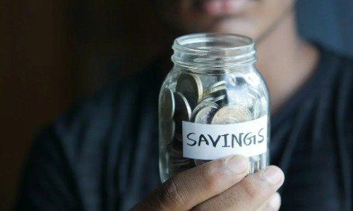 Hand holding a clear jar with coins. A strip of white tape contains the words Savings in black ink